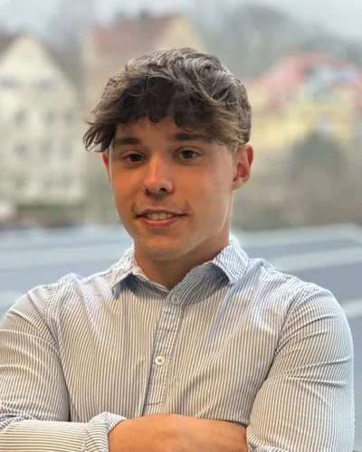 Daniel Wulf, im Halbporträt vor einer unscharfen Häuserzeile, in gestreiftem Hemd