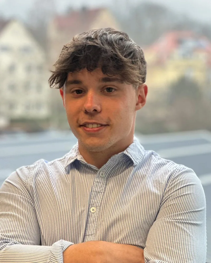 Daniel Wulf, im Halbporträt vor einer unscharfen Häuserzeile, in gestreiftem Hemd